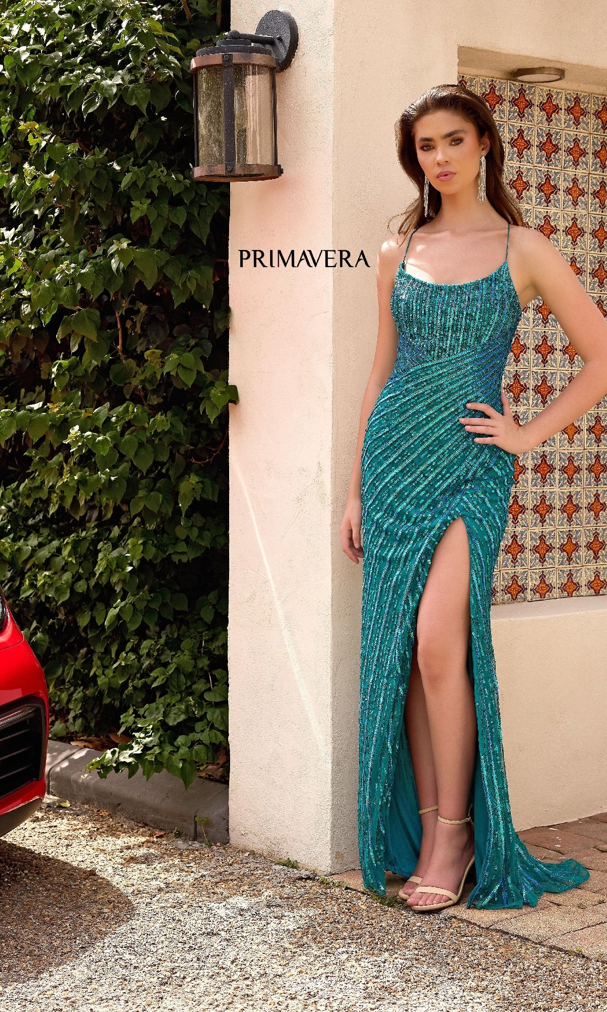 A woman in the Strappy Long Beaded Prom Dress: Primavera 4363, featuring teal sequins and a thigh-high slit, stands outdoors by a wall displaying “PRIMAVERA.”.